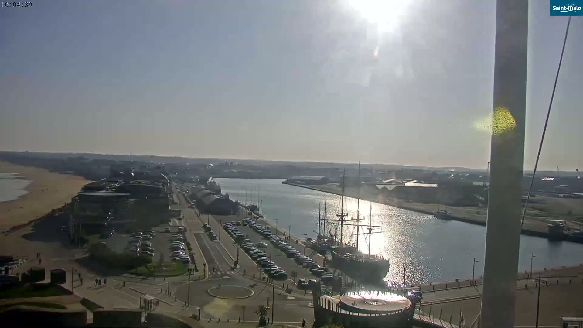 A sunny day overlooking a harbor with a large sailing ship docked, a sandy beach, and a city in the distance.