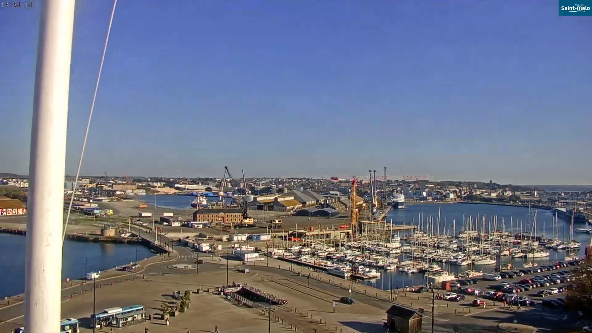 A harbor filled with numerous sailboats and yachts is shown under a clear, sunny sky, with a city and industrial area visible in the background.