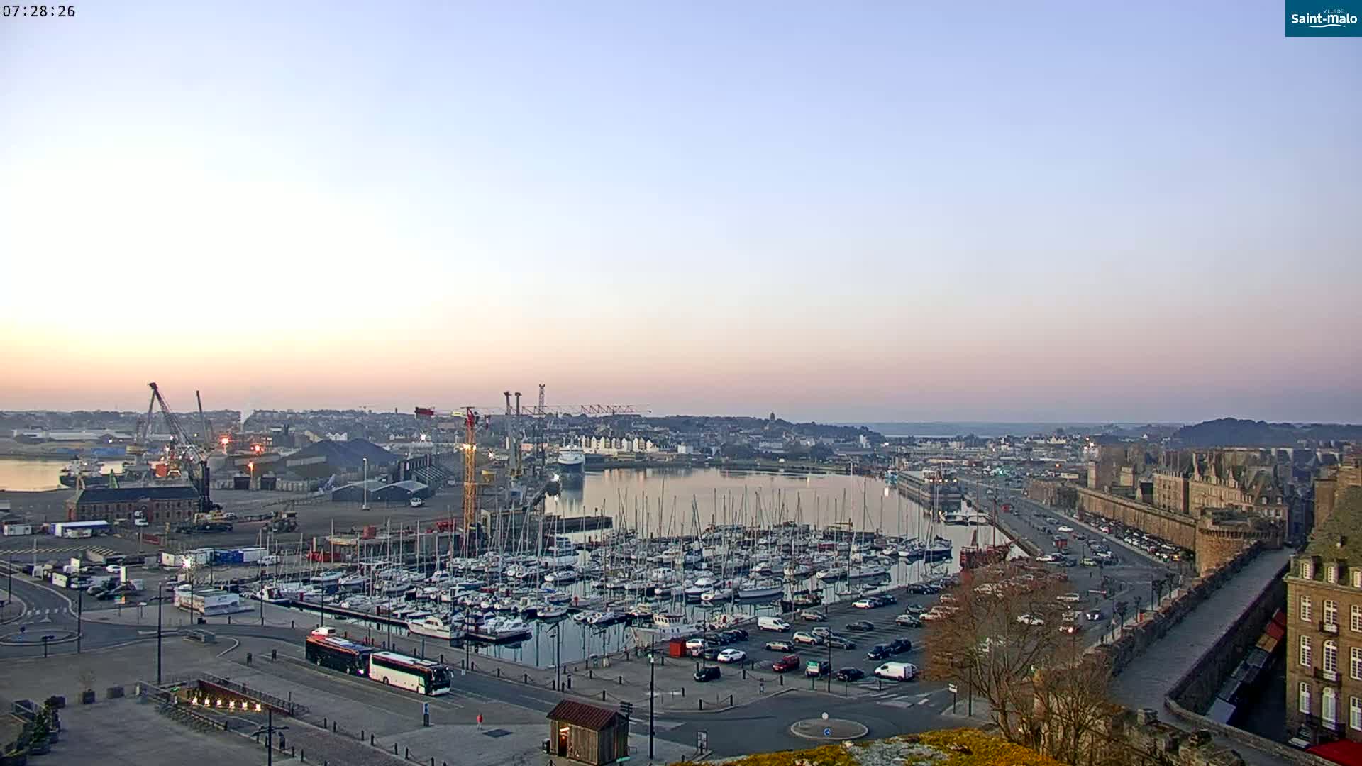 A harbor filled with numerous sailboats at sunrise, under a clear, pale-pink sky, is overlooked by a city with a fortified wall.