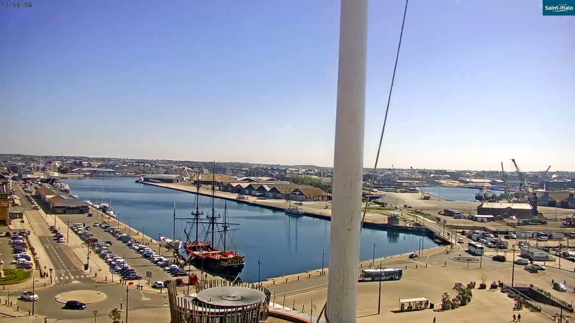 A sunny harbor scene shows a large sailing ship docked near a parking lot, with a city and additional docks visible in the background.