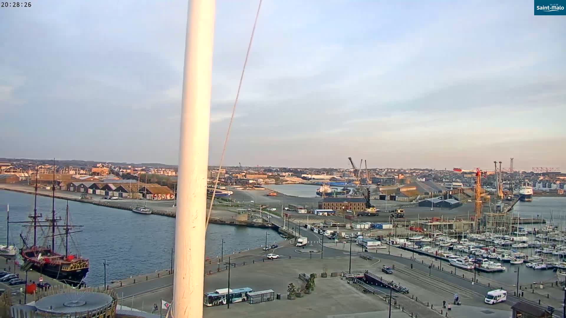 A harbor scene at dusk shows a large sailing ship, a marina filled with boats, industrial areas, and a town in the distance under a mostly clear sky.