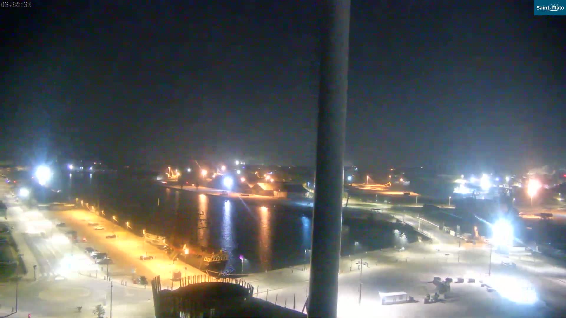 A nighttime, high-angle view of a harbor with docked boats, illuminated parking lots, and buildings along the waterfront under a clear, dark sky.