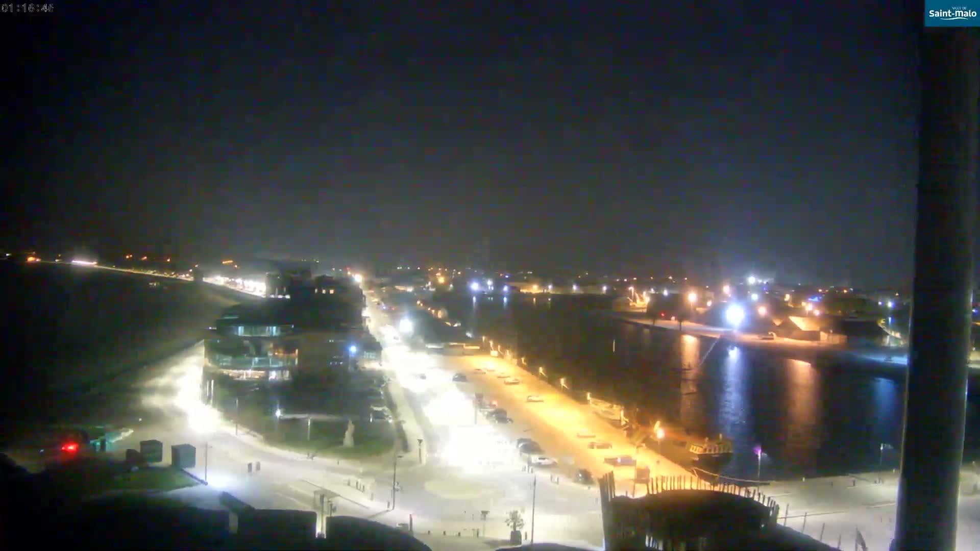 A nighttime aerial view of a waterfront city scene shows a body of water, a roadway lined with streetlights, and buildings illuminated against a dark sky.