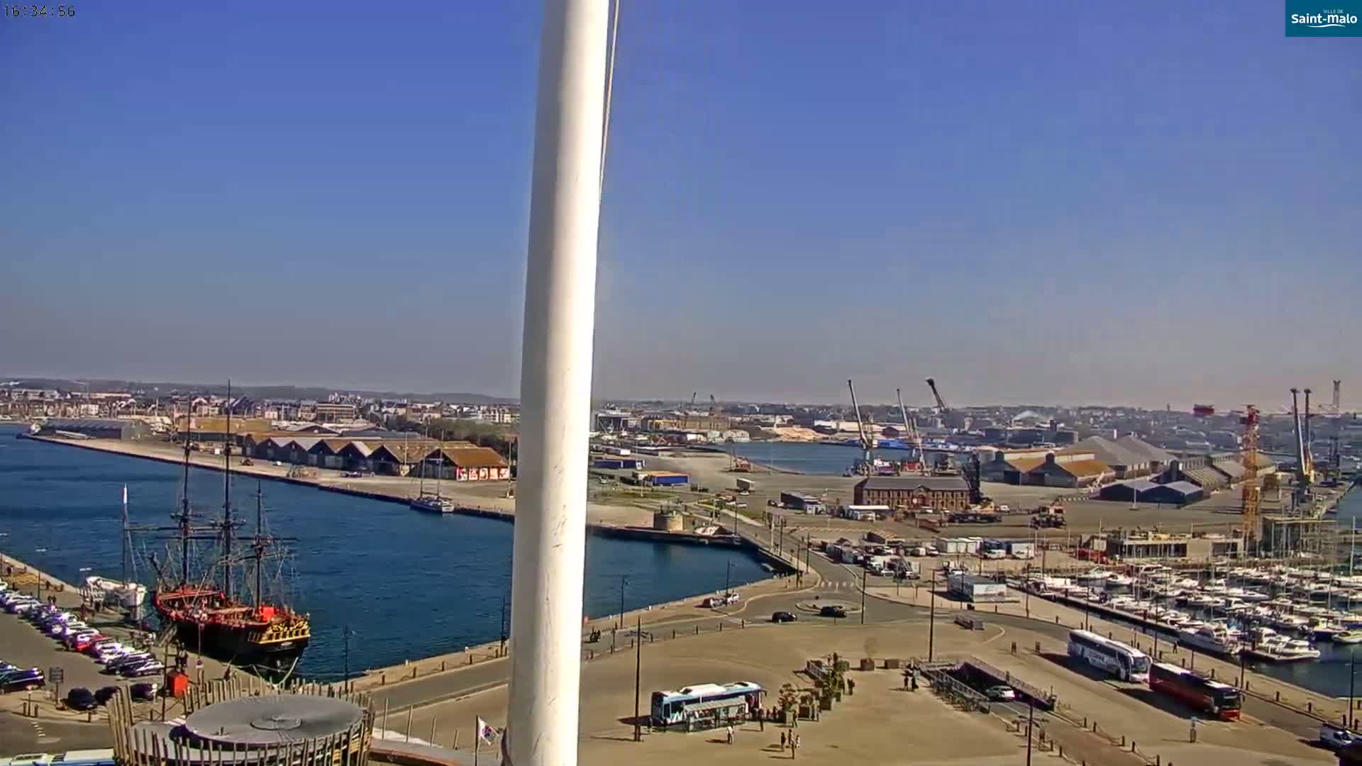 A harbor scene on a sunny day shows a large sailing ship docked near a marina filled with smaller boats, with a city and industrial area in the background.