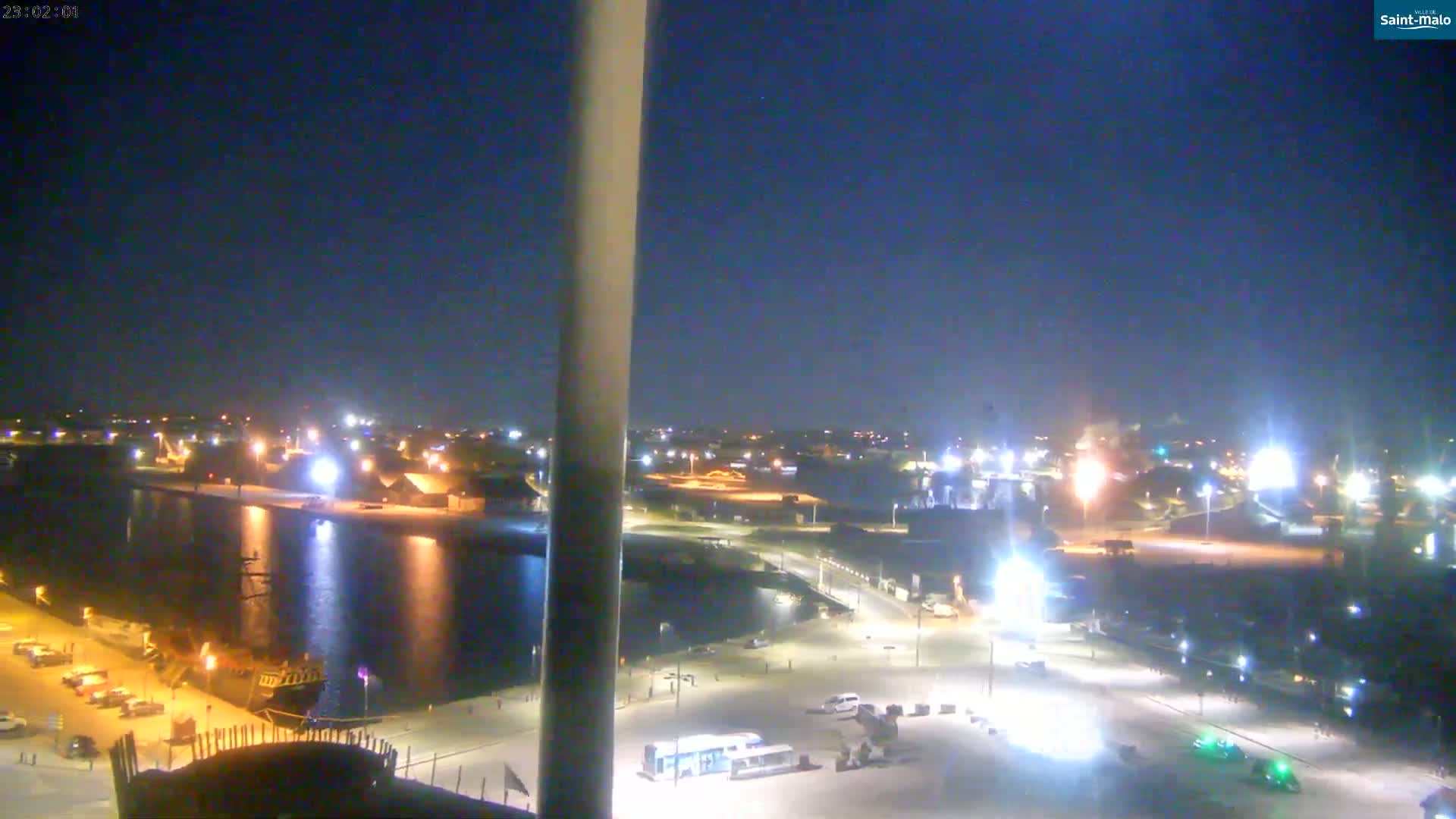 A nighttime view of a harbor with a docked ship, illuminated buildings, and parked vehicles under a clear, dark sky.