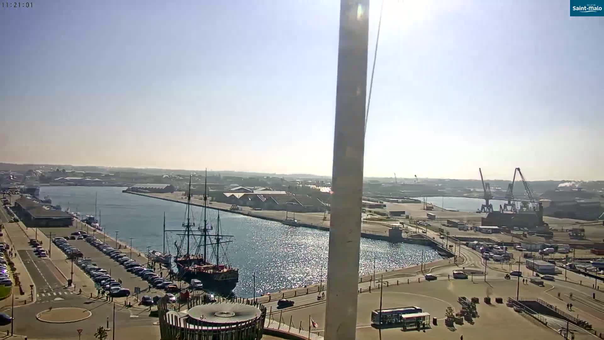 A sunny day overlooking a harbor with a large sailing ship docked near a parking lot and industrial area in the distance.
