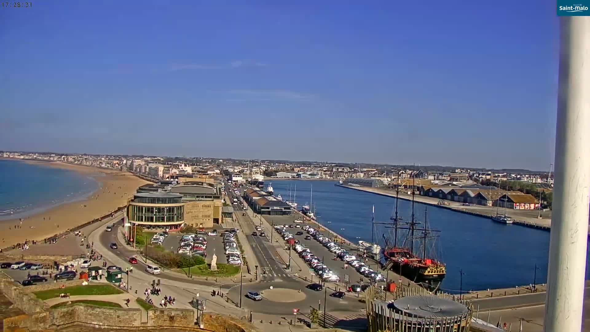 A sunny, high-angle view shows a harbor with a large sailing ship, a sandy beach, and a coastal town extending into the distance.