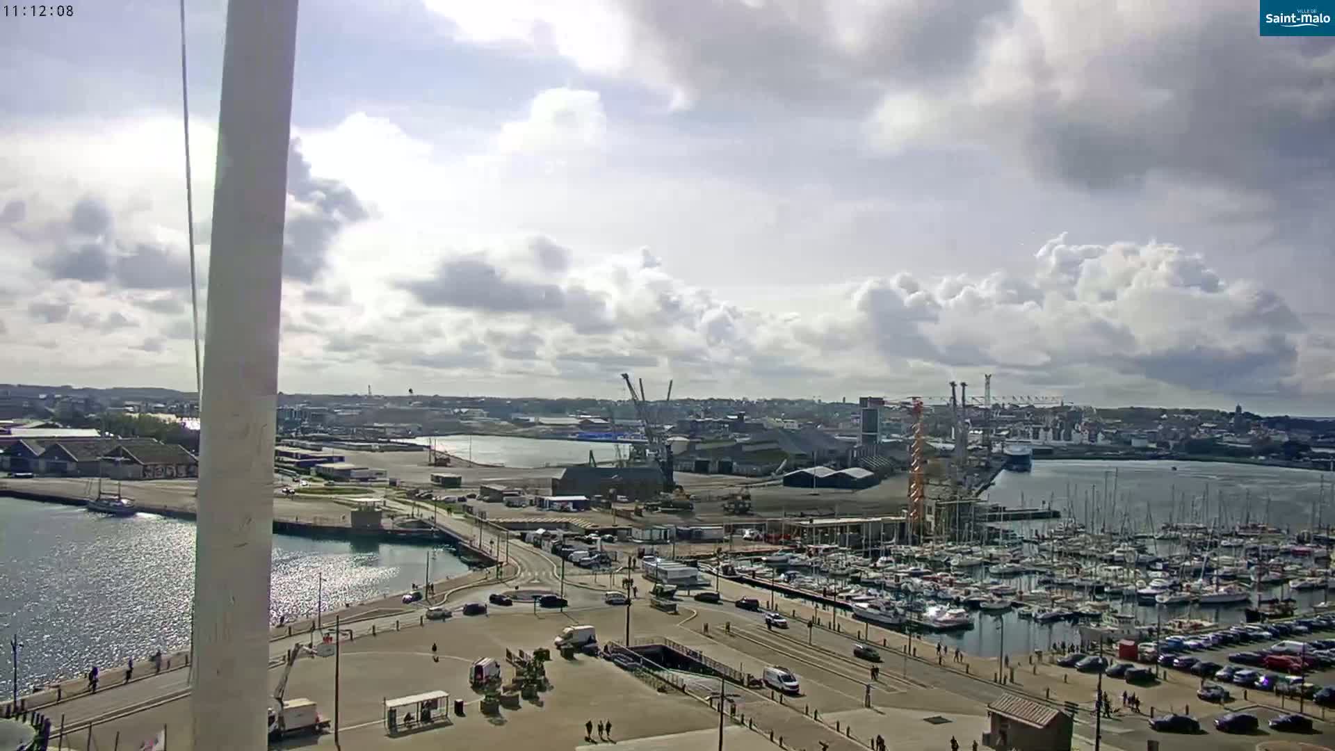 A partly cloudy day reveals a harbor filled with sailboats and a city with industrial areas and cranes, viewed from a high vantage point.