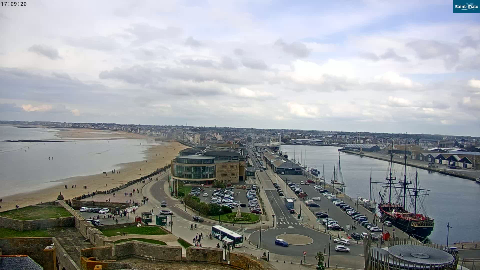An aerial view shows a coastal city under a partly cloudy sky, featuring a sandy beach, a harbor with a large ship, and buildings lining a waterfront road.