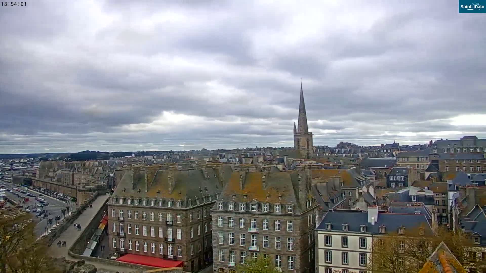 An elevated view of a European city under a cloudy sky shows numerous buildings with dark gray slate roofs, and a tall church spire in the distance.