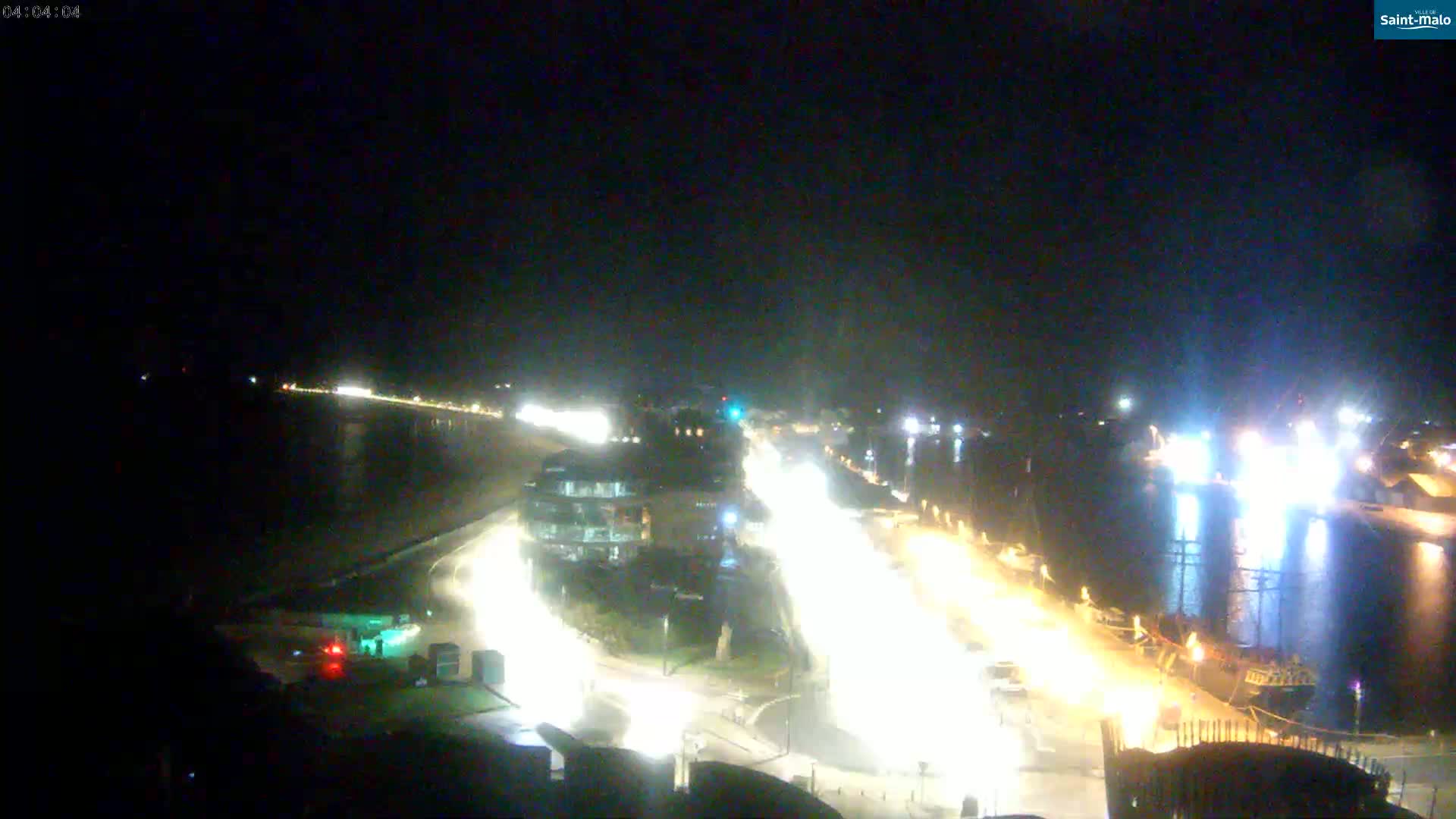 A nighttime aerial view shows a waterfront area with brightly lit roads and buildings, a body of water with a docked ship, and clear skies.