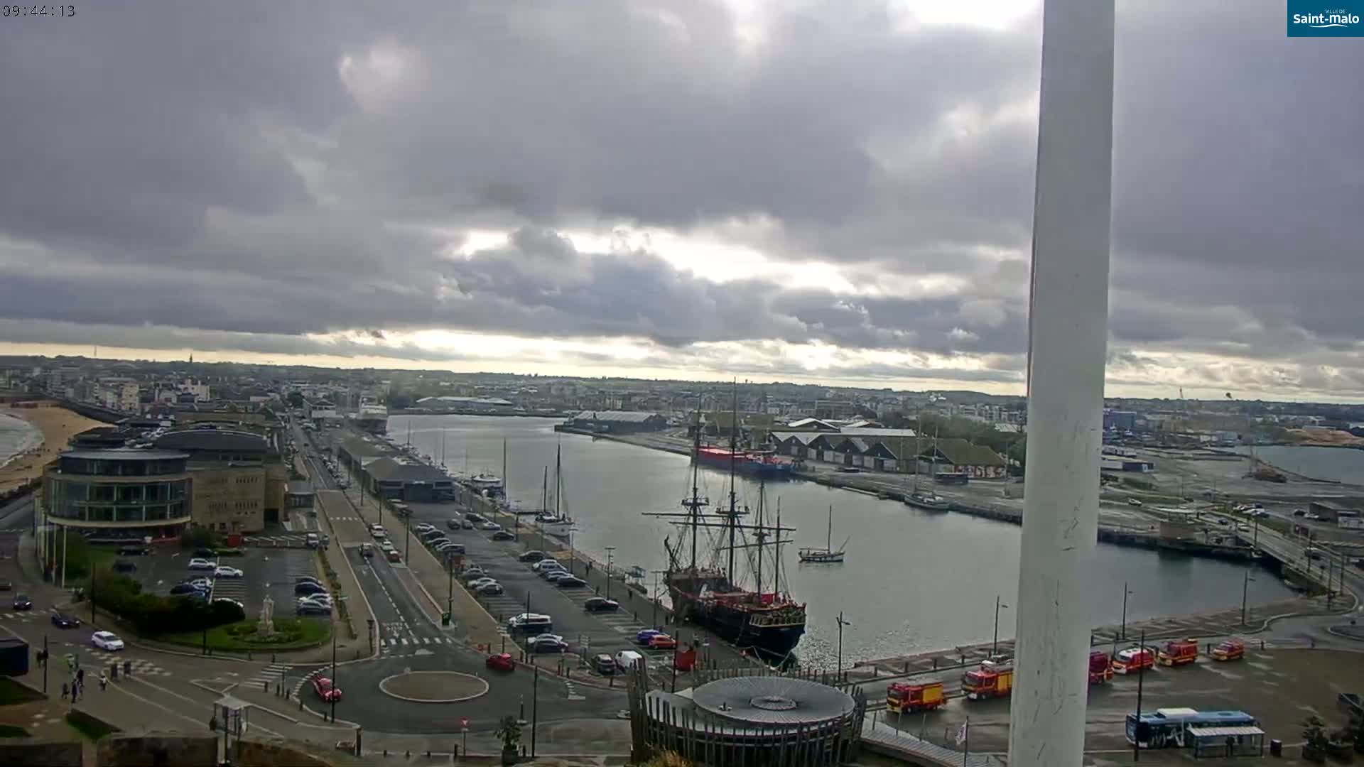 A harbor scene under a cloudy sky shows a large sailing ship docked near a city, with cars parked along the waterfront and emergency vehicles visible.