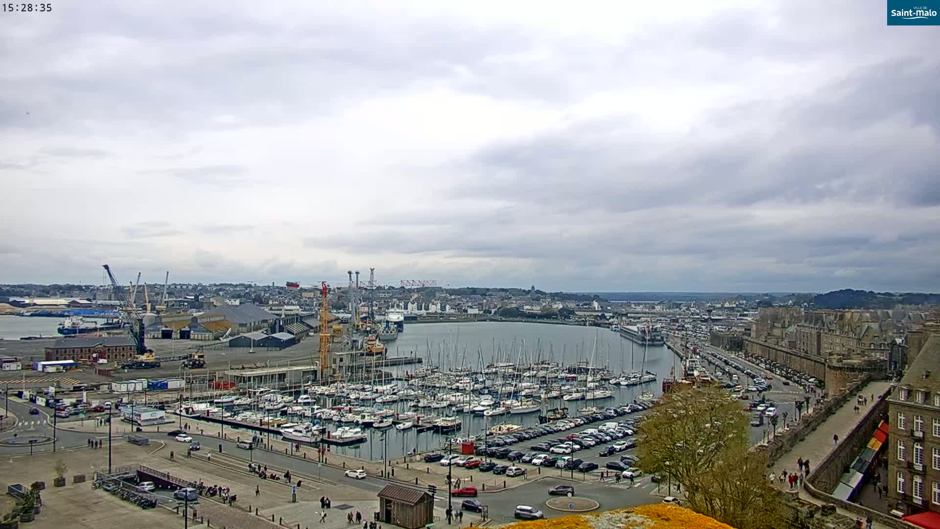 A harbor filled with numerous sailboats and yachts under a mostly cloudy sky, with a city and port facilities visible in the background.