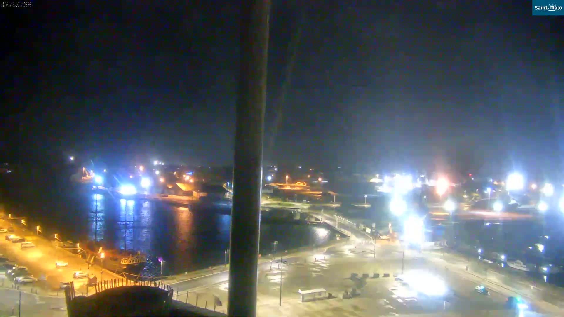 A nighttime, brightly lit harbor scene shows several boats and vehicles near a waterfront with a bridge visible in the distance.