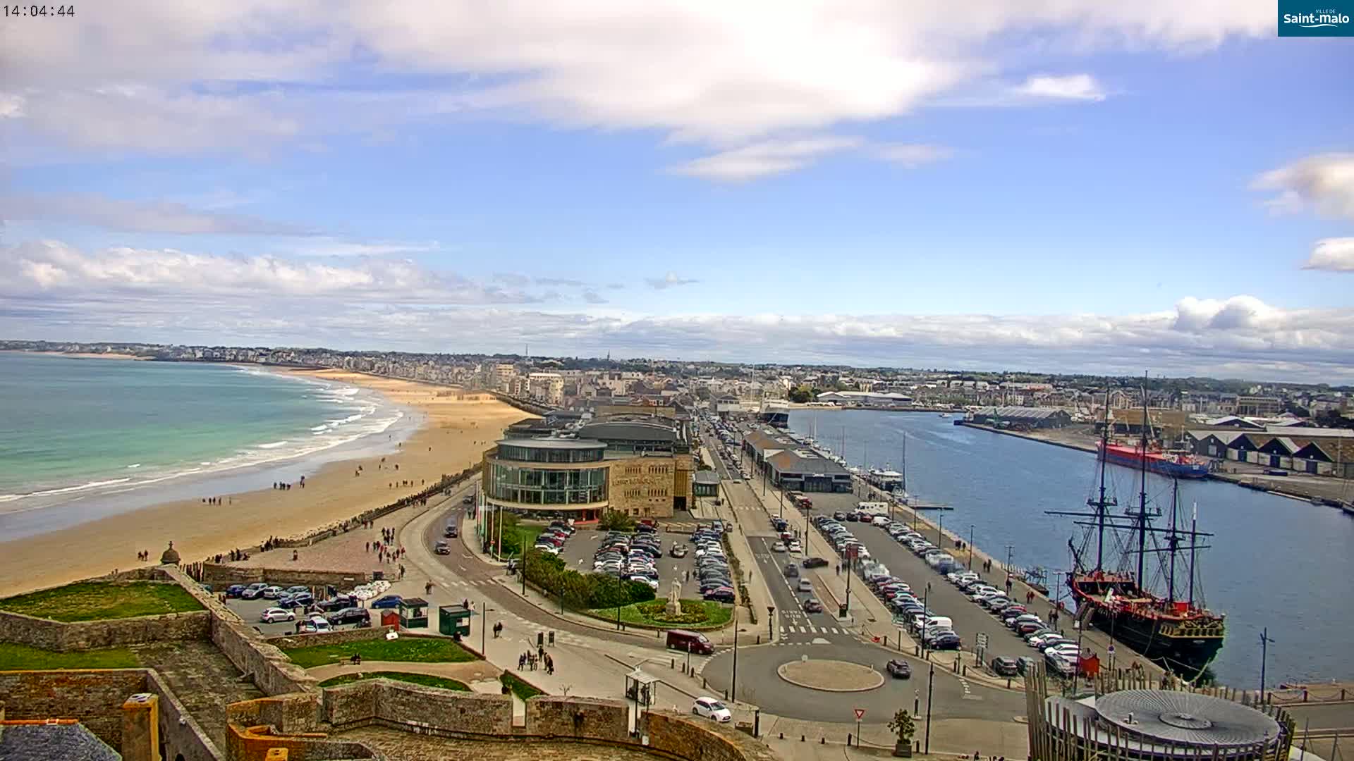 A coastal city with a sandy beach, a harbor containing a large sailing ship, and a partly cloudy sky is viewed from an elevated position.