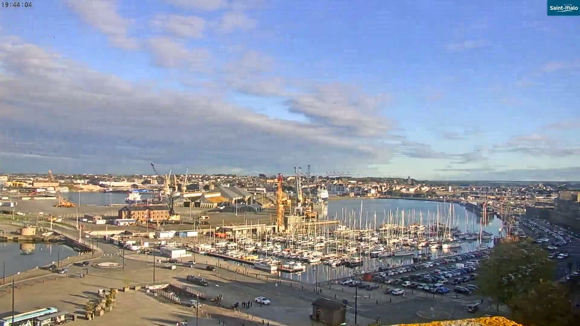A partly cloudy day reveals a harbor filled with numerous sailboats and other vessels, alongside a city and industrial areas.