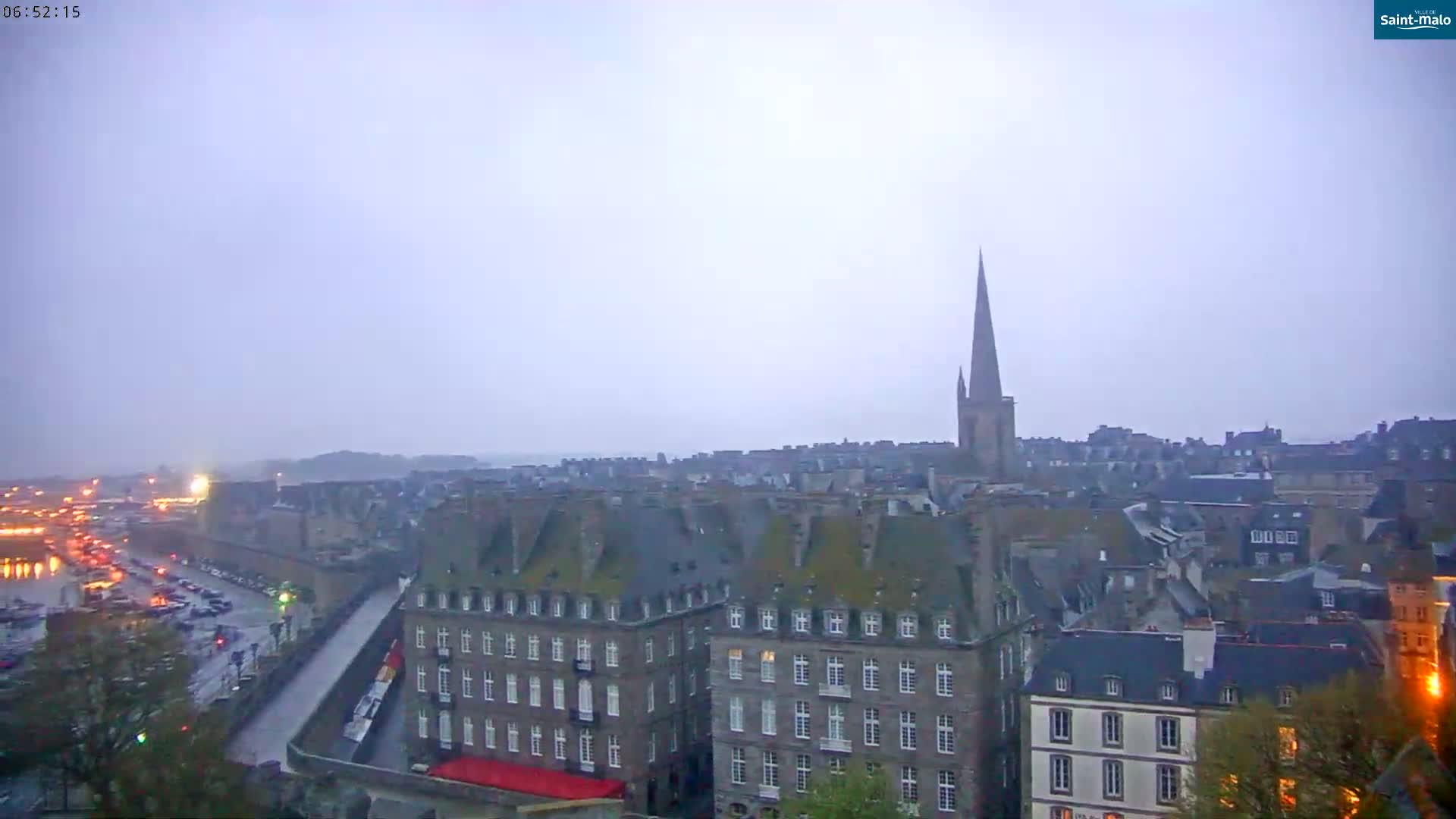 A hazy, overcast view of a city with a tall church spire in the distance, overlooking buildings and a waterfront with traffic.