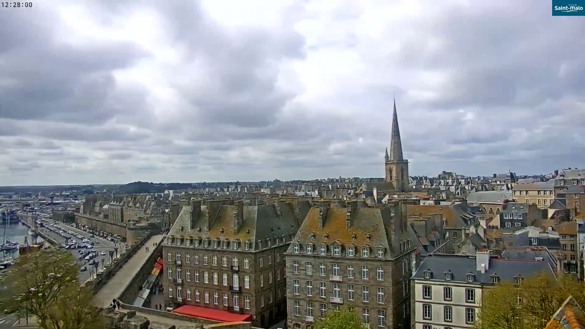 A sprawling cityscape with numerous gray-roofed buildings, a prominent church spire, and a harbor visible in the distance under a mostly cloudy sky.