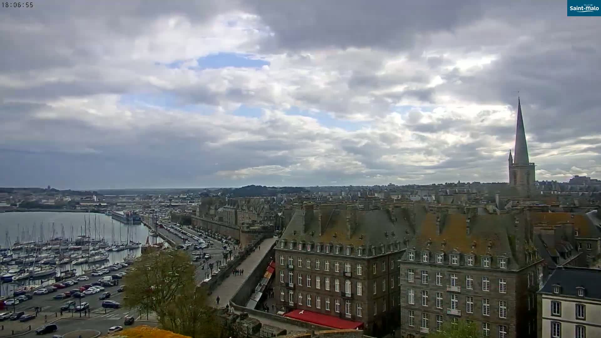 A partly cloudy sky overlooks a coastal city with a harbor full of sailboats and a tall church spire.