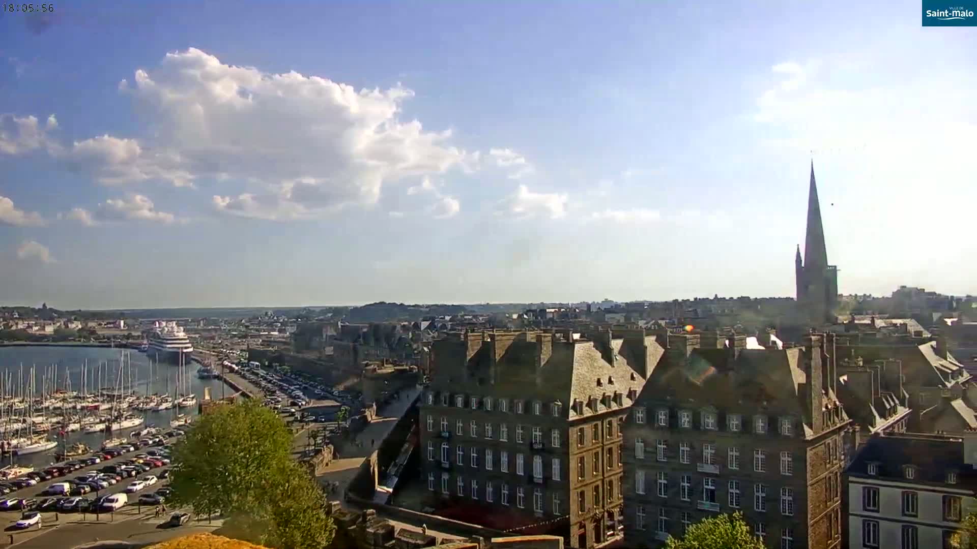 A harbor filled with sailboats and a large cruise ship is visible, along with a city featuring buildings with steeply pitched roofs under a partly sunny sky.