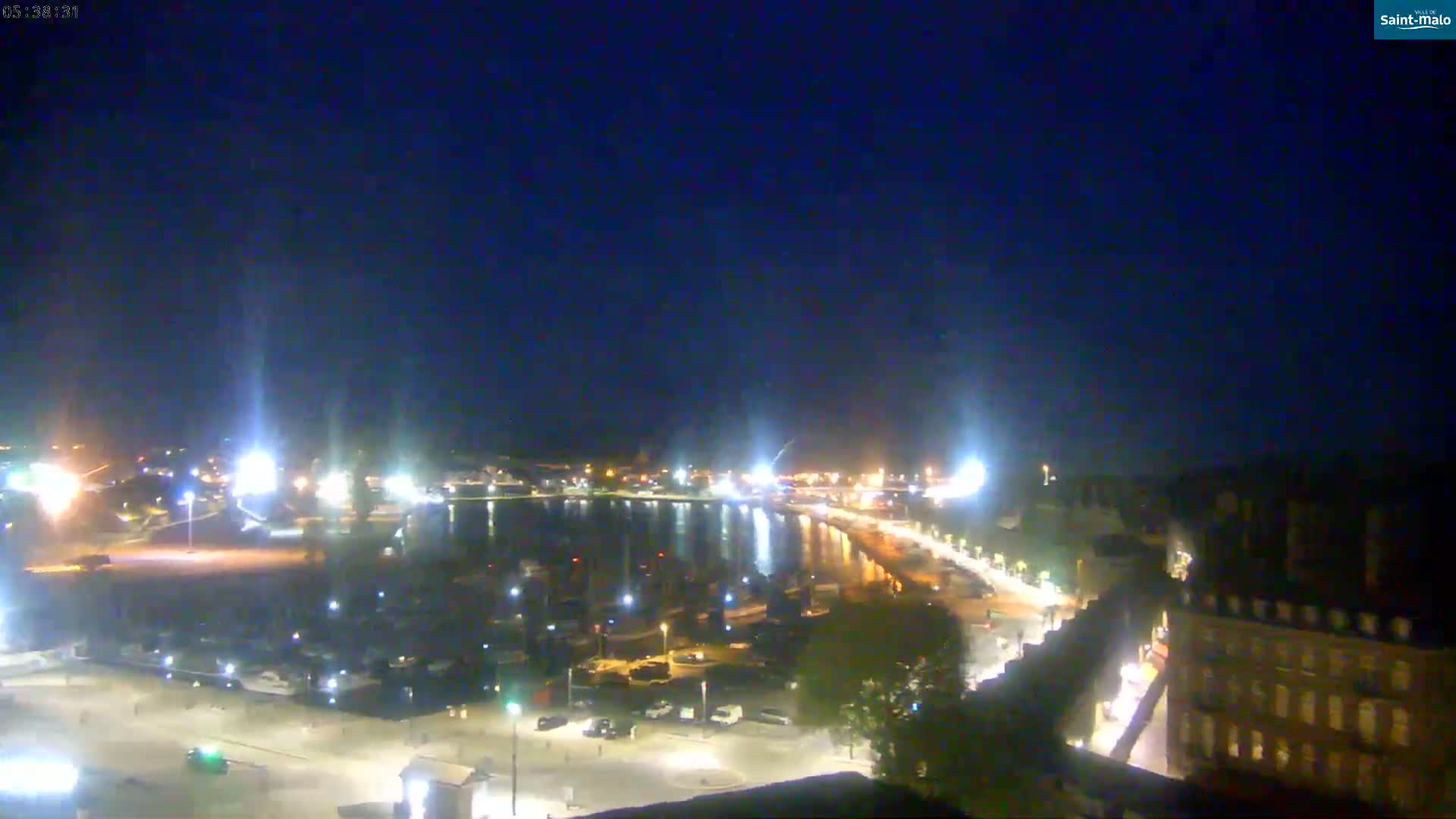 A nighttime view of a harbor filled with boats, a waterfront road with lights, and buildings along the shore under a clear, dark sky.