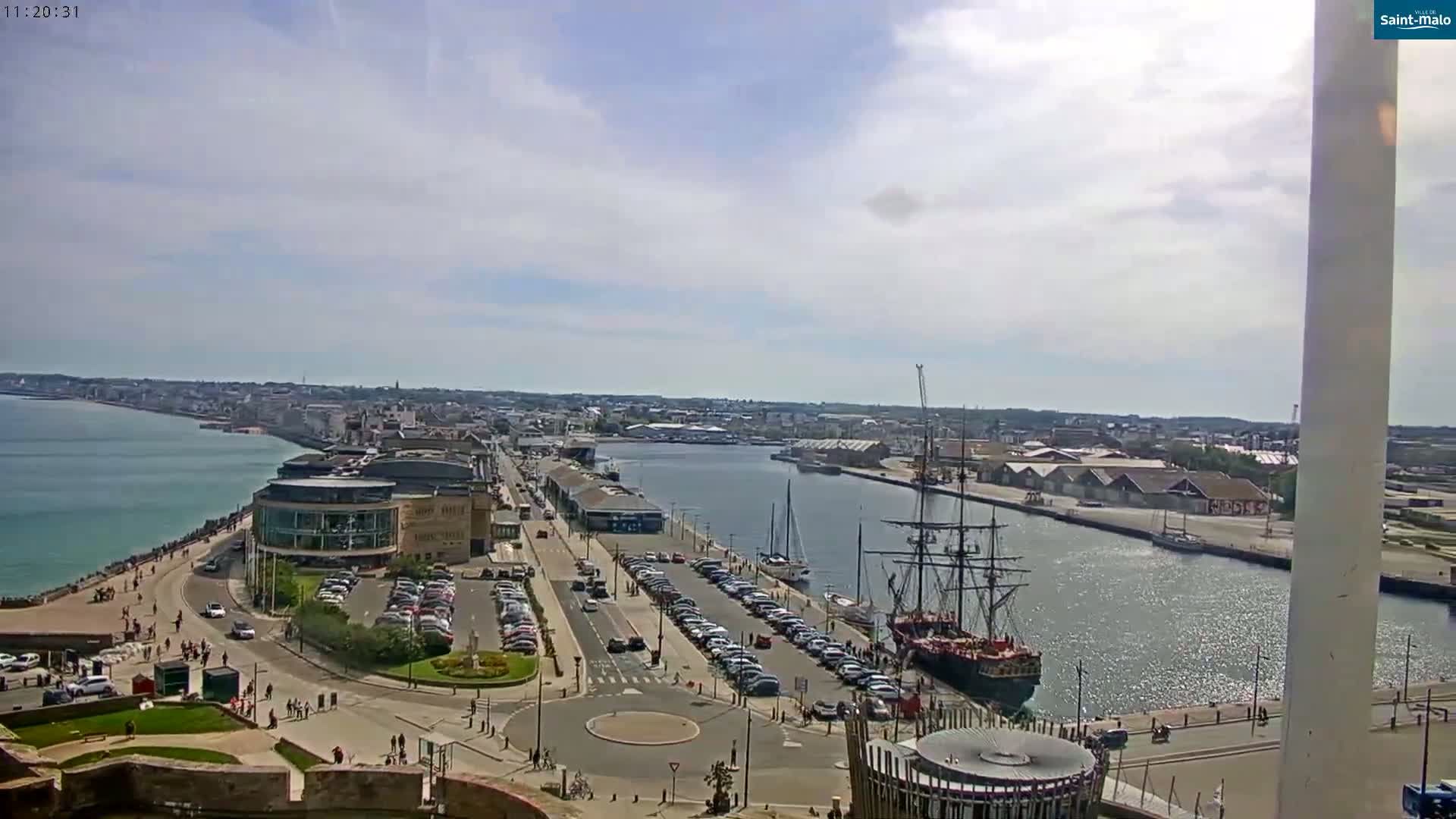 A high-angle view shows a harbor town on a partly cloudy day, with a large sailing ship docked near a waterfront area with cars parked along the road.