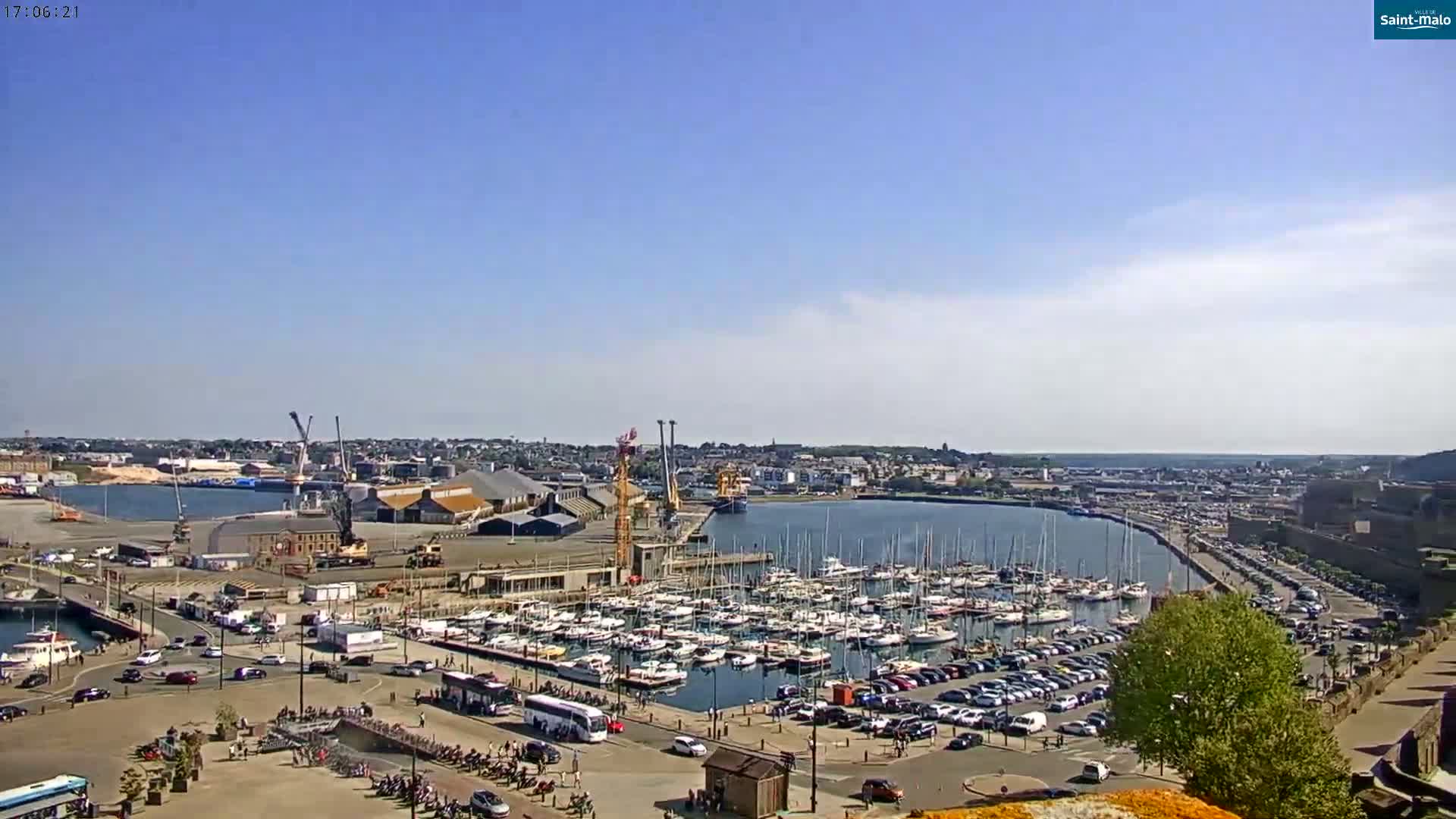 A sunny day reveals a harbor filled with numerous boats and yachts, surrounded by buildings, parking lots, and a roadway, with a city extending beyond.