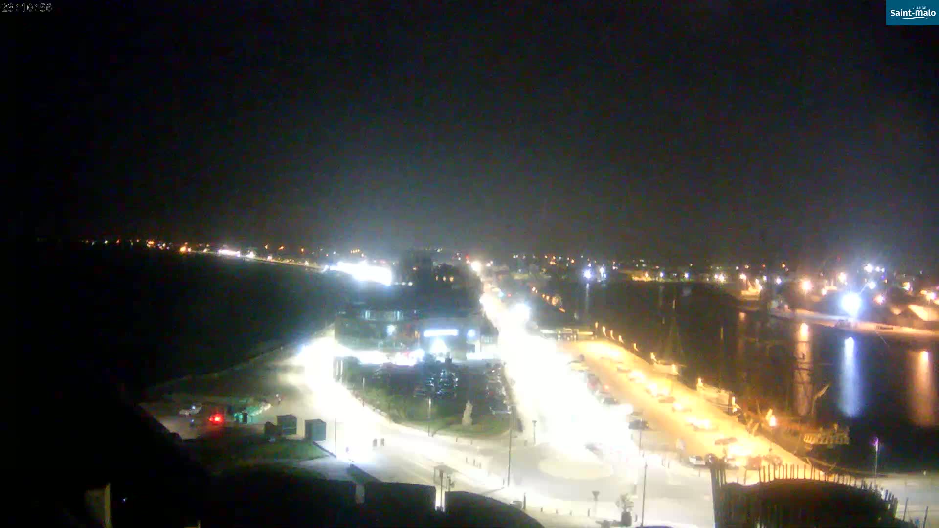A nighttime, high-angle view shows brightly lit roads and a harbor with boats, all set against a dark sky.