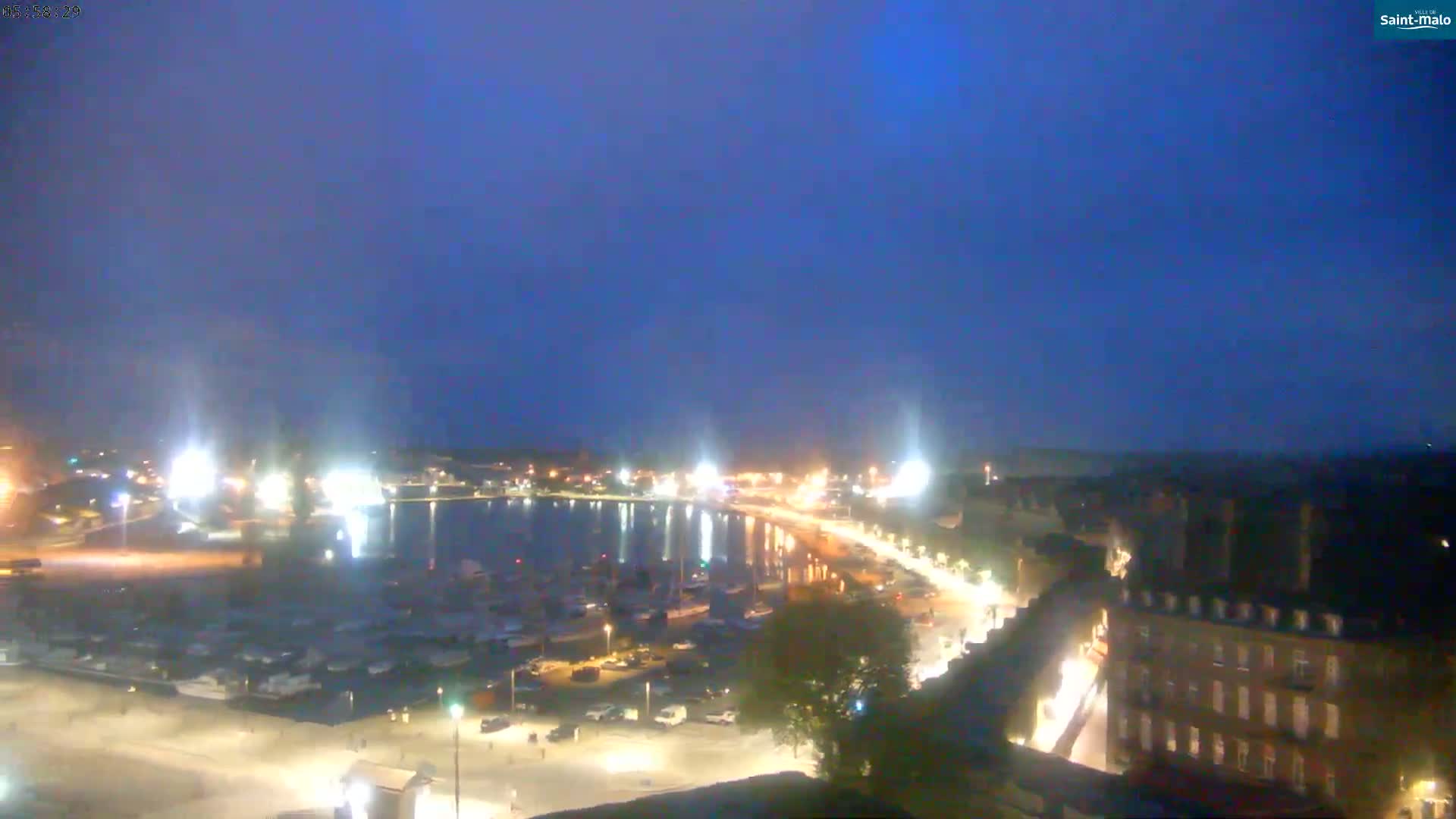 A nighttime view of a harbor filled with boats, a waterfront road lined with buildings, and a mostly clear, dark blue sky.