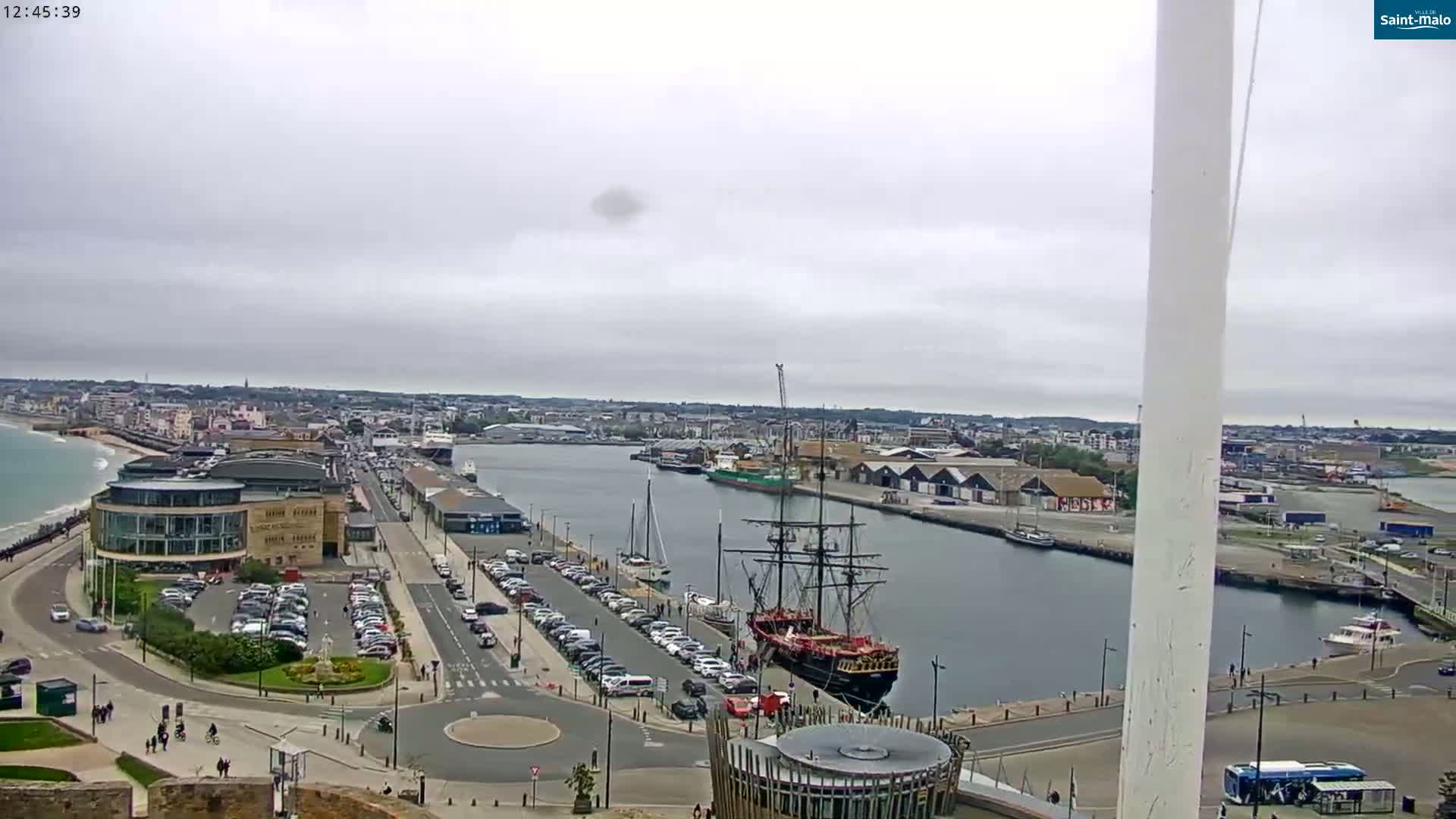 A large sailing ship is docked in a harbor, with overcast skies and a city visible in the background, numerous cars parked along the waterfront, and a beach visible to the left.
