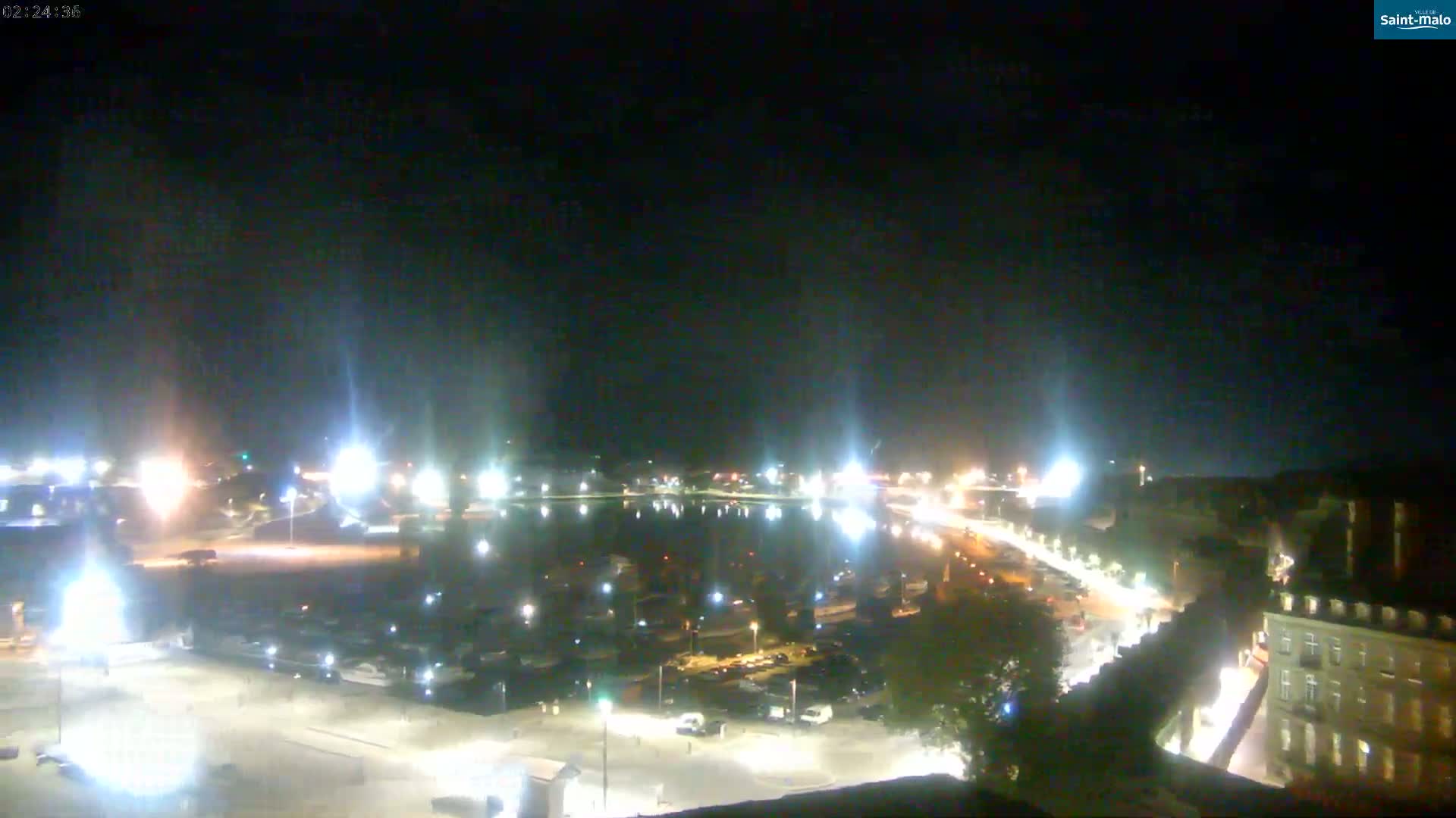 A nighttime aerial view of a harbor, illuminated by streetlights and building lights, with a calm body of water reflecting the lights.