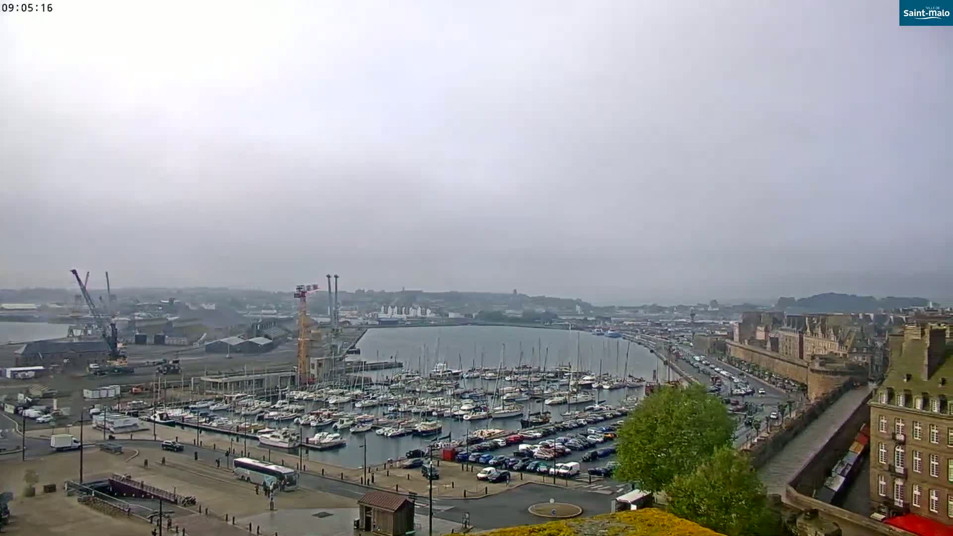 A harbor filled with numerous sailboats and motorboats is visible under an overcast sky, with a city and port infrastructure in the background.