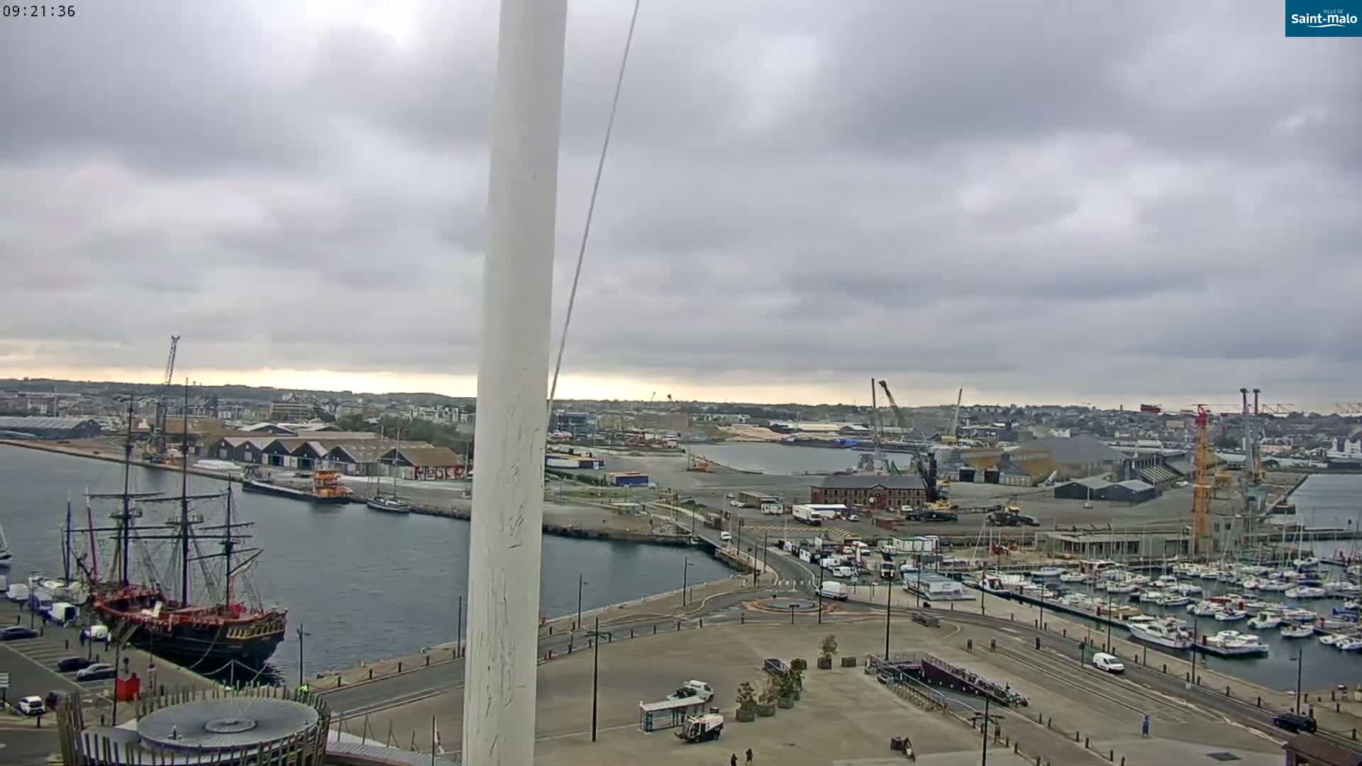 A large sailing ship is docked in a harbor under an overcast sky, with numerous other boats and a cityscape in the background.