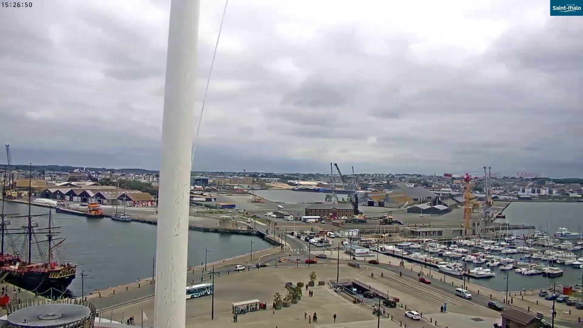 An overcast day reveals a harbor scene with a large sailing ship, numerous docked boats, industrial areas, and a tall white structure in the foreground.