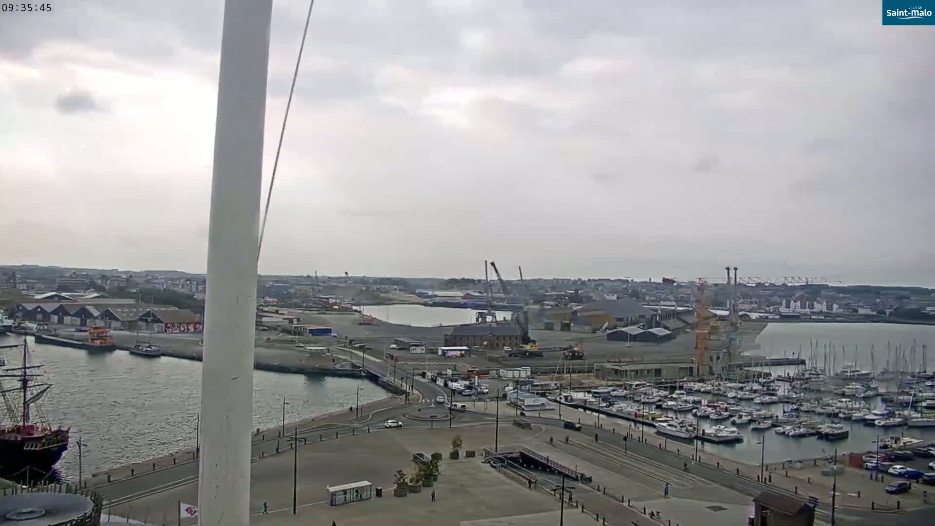 A high-angle, long shot shows a harbor with numerous boats and ships on a cloudy day.