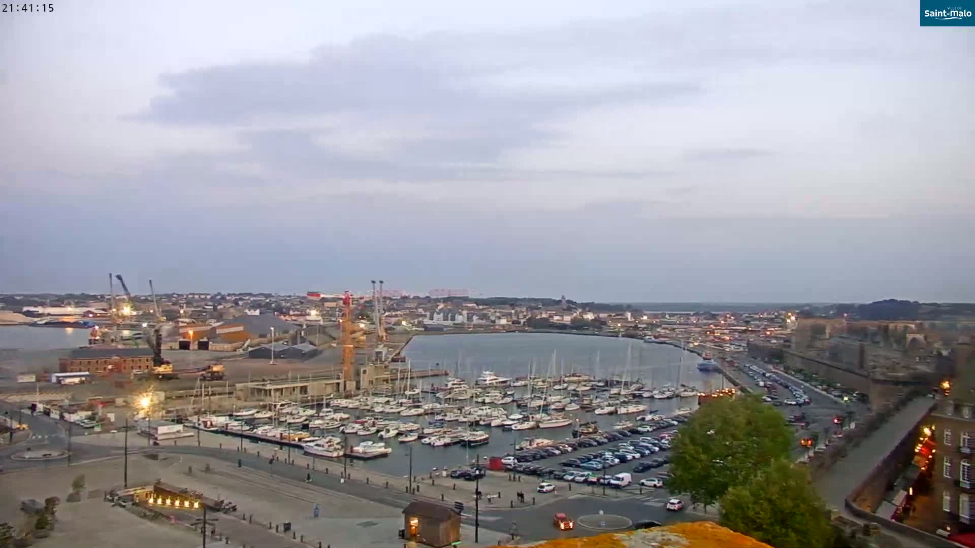 A harbor filled with sailboats and yachts is situated next to a city under a cloudy, twilight sky.