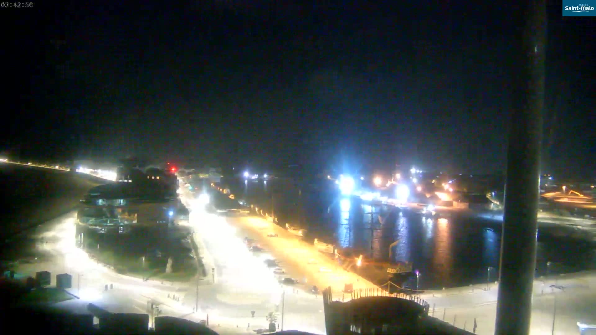 A nighttime view of a harbor and city area with brightly lit roads and buildings near a body of water where ships are docked.
