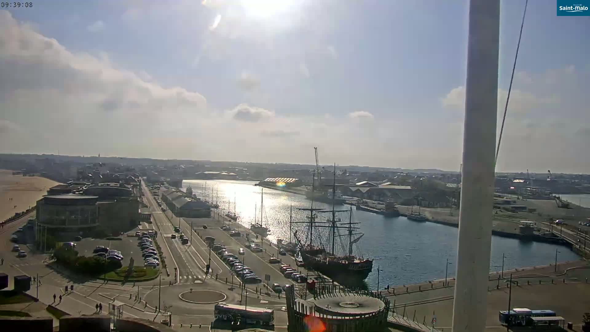 A high-angle view shows a harbor with a large sailing ship docked, a city skyline, and partly sunny skies.