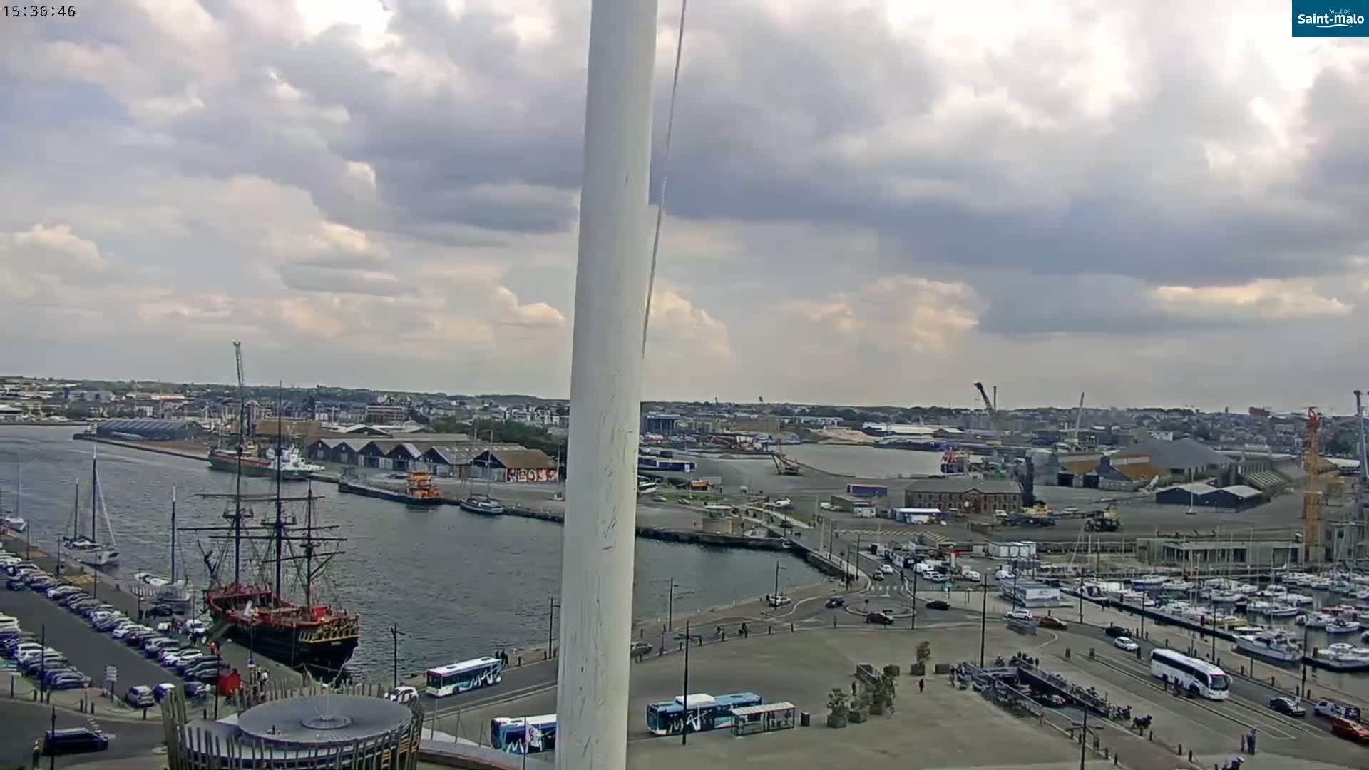 A partly cloudy day overlooks a harbor with a large sailing ship docked near a city, where cars are parked along the waterfront and buses are driving near a tall, white structure.