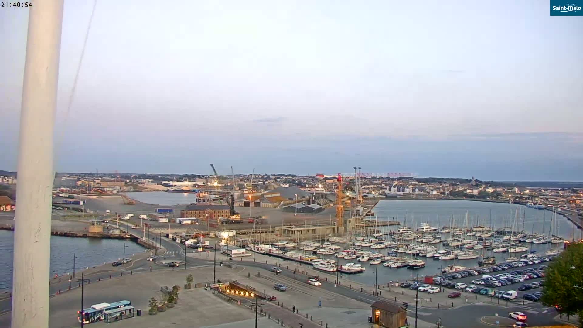 A harbor filled with sailboats and yachts is viewed from an elevated position at dusk under a clear, light sky.
