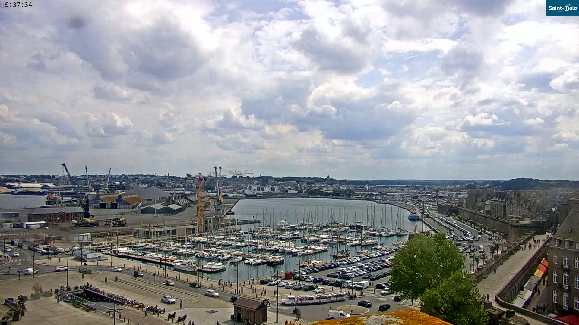 A partly cloudy day reveals a harbor filled with sailboats and yachts, adjacent to a city with buildings and a stone wall.