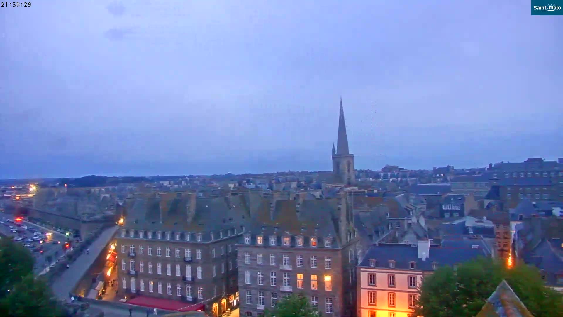 A twilight view shows a city with many buildings and a tall spire, under a clear, dusky sky.