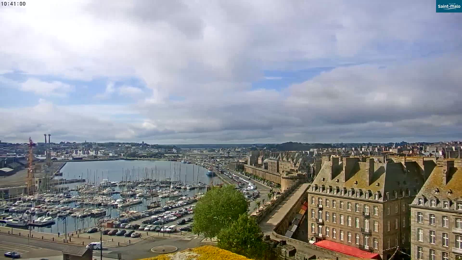 A partly cloudy day reveals a harbor filled with sailboats and yachts, adjacent to a city with stone buildings, a walled area, and a parking lot.