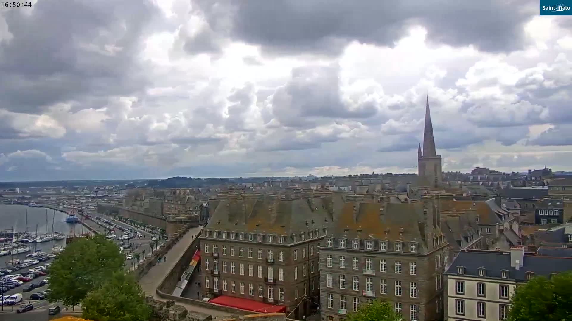 A partly cloudy sky overlooks a coastal city with a harbor full of boats and numerous stone buildings, including a tall church spire.