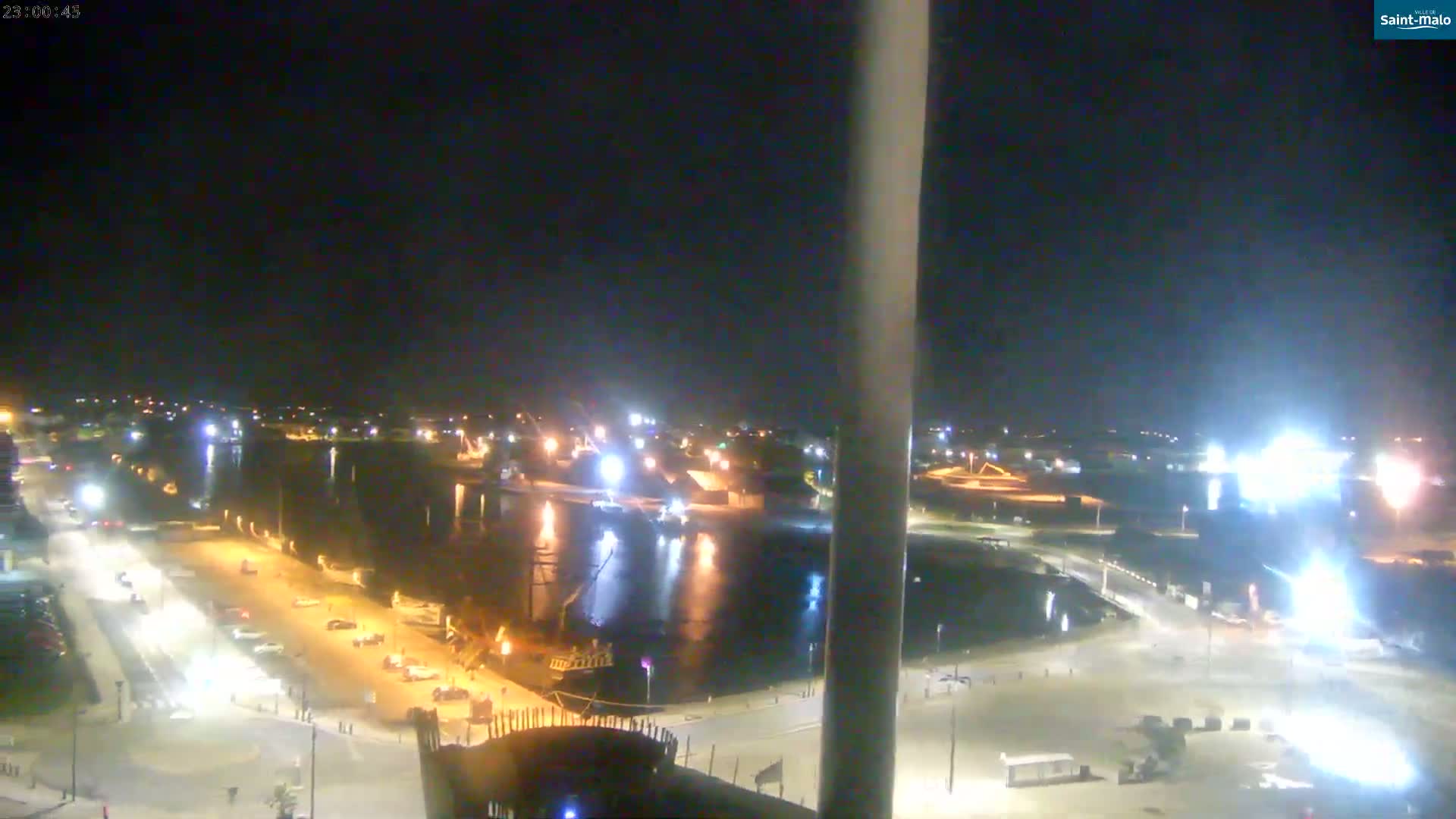 A nighttime aerial view shows a harbor with boats and docked ships, surrounding roads with parked cars, and brightly lit industrial areas.