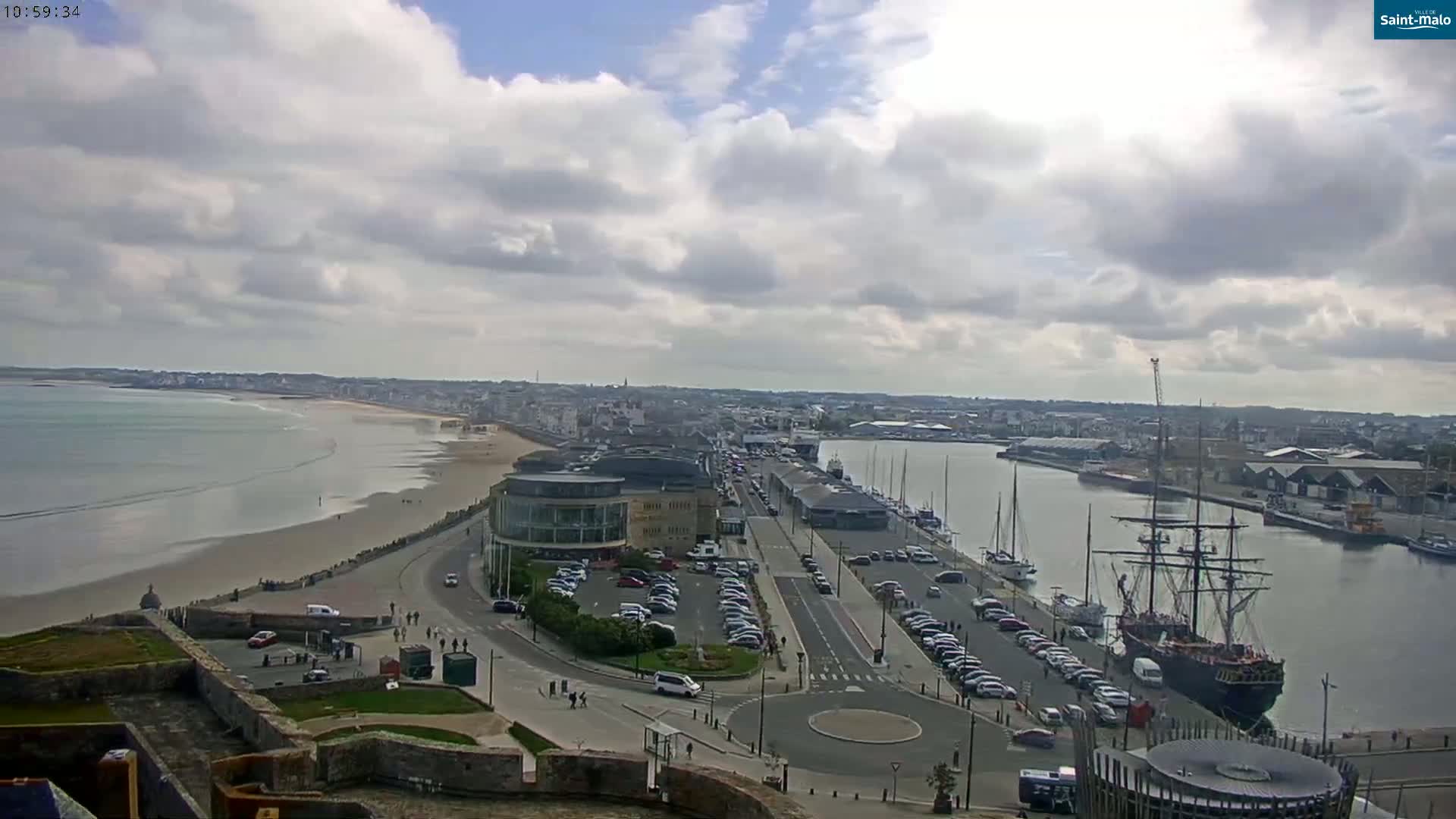 A coastal city with a harbor featuring a large sailing ship is viewed from a high vantage point on a partly cloudy day.