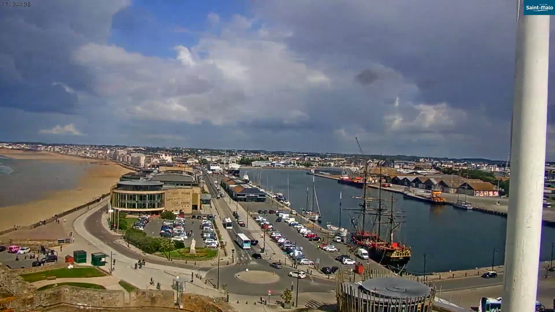 A partly cloudy day overlooks a coastal city with a harbor containing a large sailing ship, a sandy beach, and numerous parked cars.