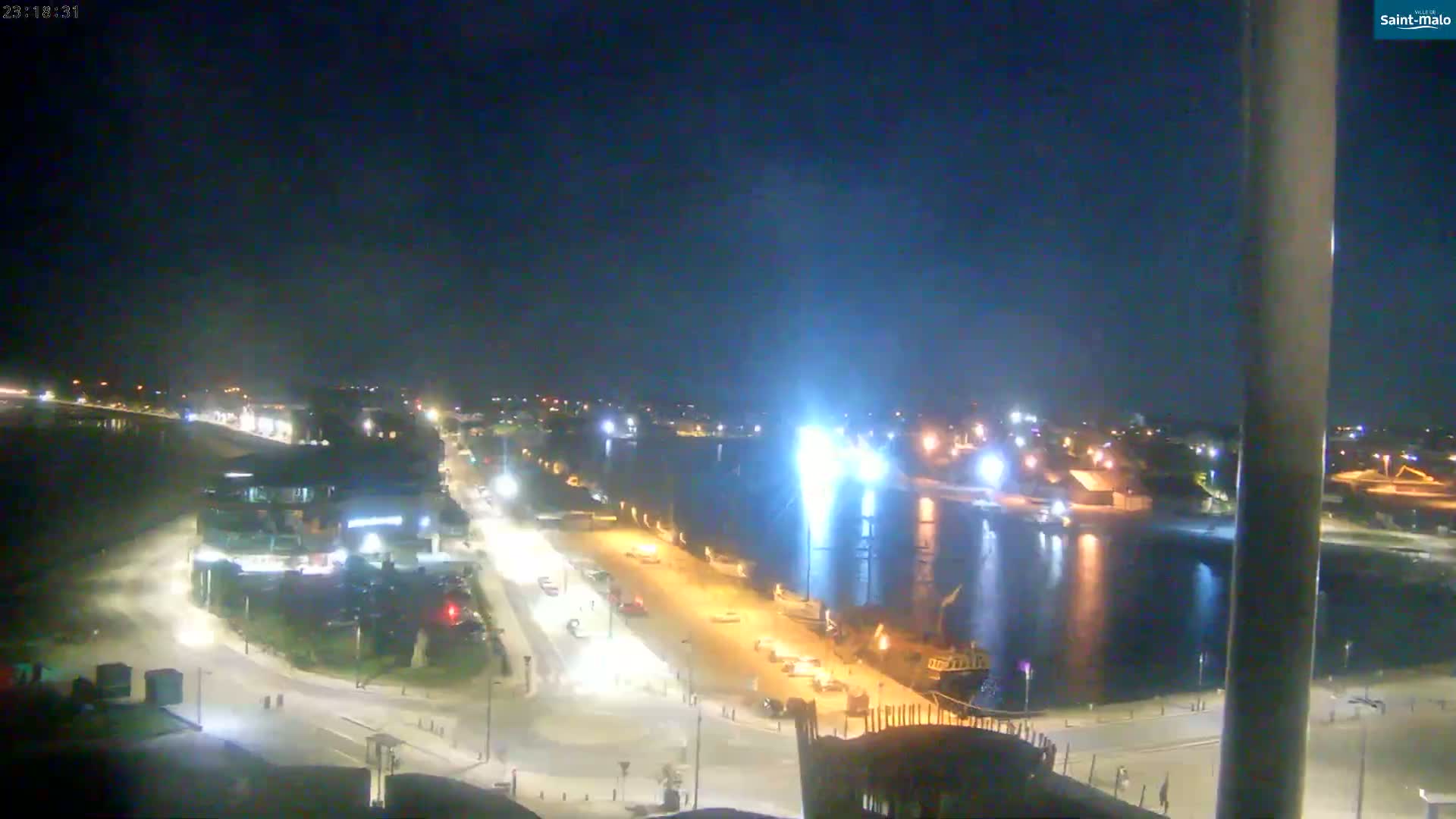 A nighttime, high-angle view shows a waterfront area with a calm body of water, illuminated streets, and buildings along the shore.