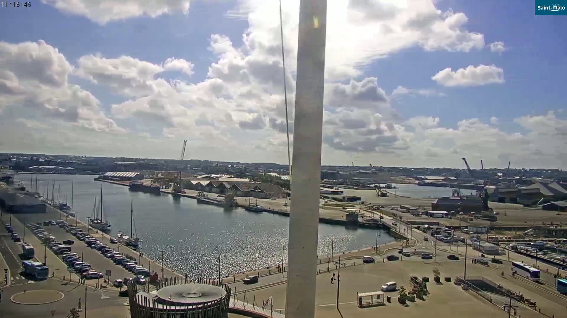 A sunny day with partly cloudy skies overlooks a harbor filled with boats, industrial buildings, and a parking lot.