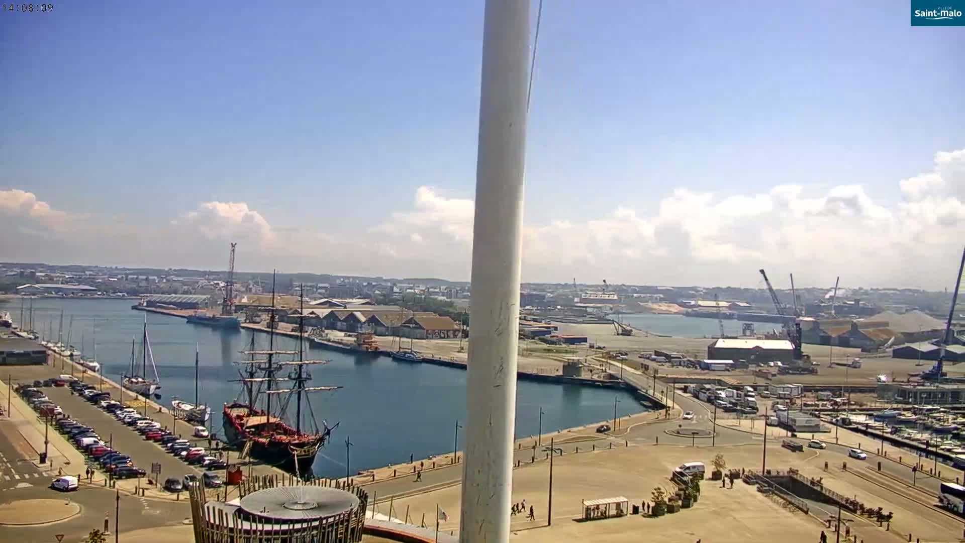 A harbor scene on a partly sunny day shows a large sailing ship docked near a parking lot and other boats, with industrial areas and cranes in the background.